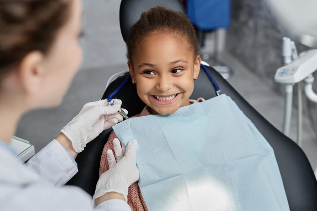 A smiling young girl sitting in a dental chair as a dentist in gloves prepares a bib.