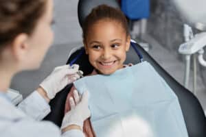 A smiling young girl sitting in a dental chair as a dentist in gloves prepares a bib.