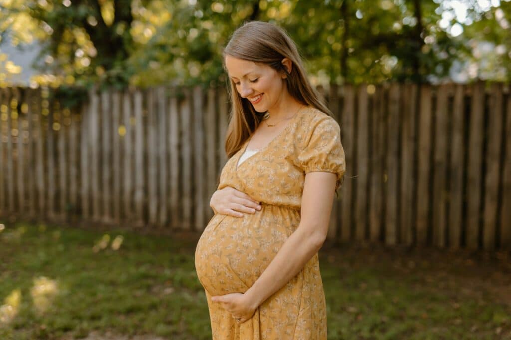 A pregnant woman smiles down at her baby bump outdoors, preparing for the future and her child’s first pediatric dental visit.