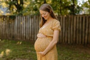 A pregnant woman smiles down at her baby bump outdoors, preparing for the future and her child’s first pediatric dental visit.