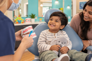 A dentist demonstrates proper brushing on a model for a smiling young boy holding a toy elephant during his first pediatric dental visit, while his mother watches.