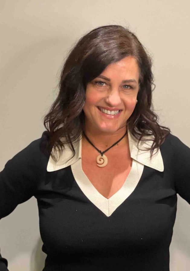 A woman with dark hair smiles, wearing a black shirt with a white collar and a spiral pendant necklace.