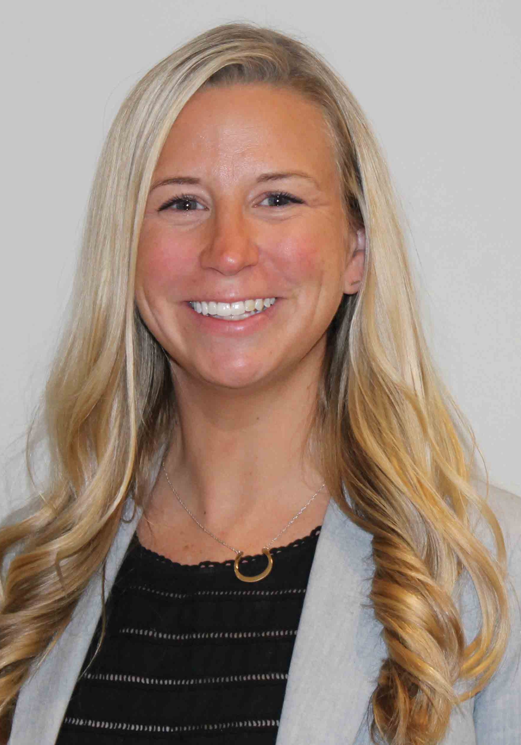 A smiling blonde woman wearing a light-colored blazer over a black top and a gold necklace.