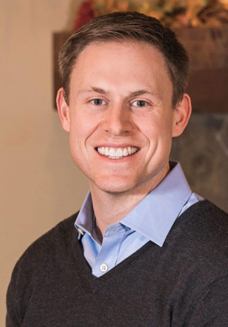 Headshot of a smiling man wearing a light blue collared shirt under a dark v-neck sweater.
