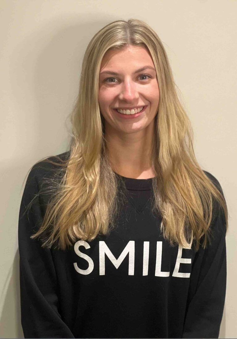 A woman with long blonde hair smiling, wearing a black sweatshirt with the word “SMILE” printed on the front.