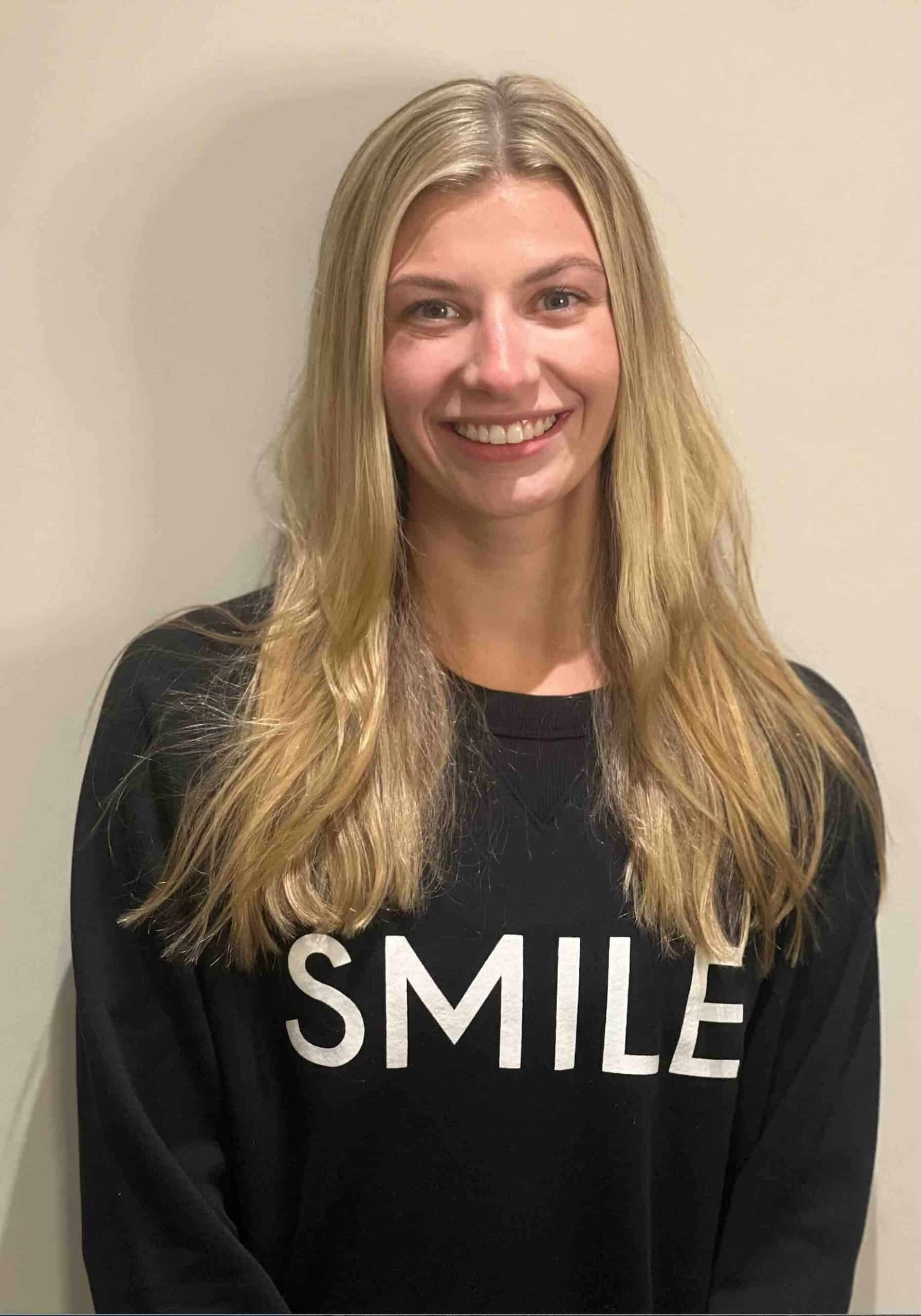 A woman with long blonde hair smiling, wearing a black sweatshirt with the word “SMILE” printed on the front.