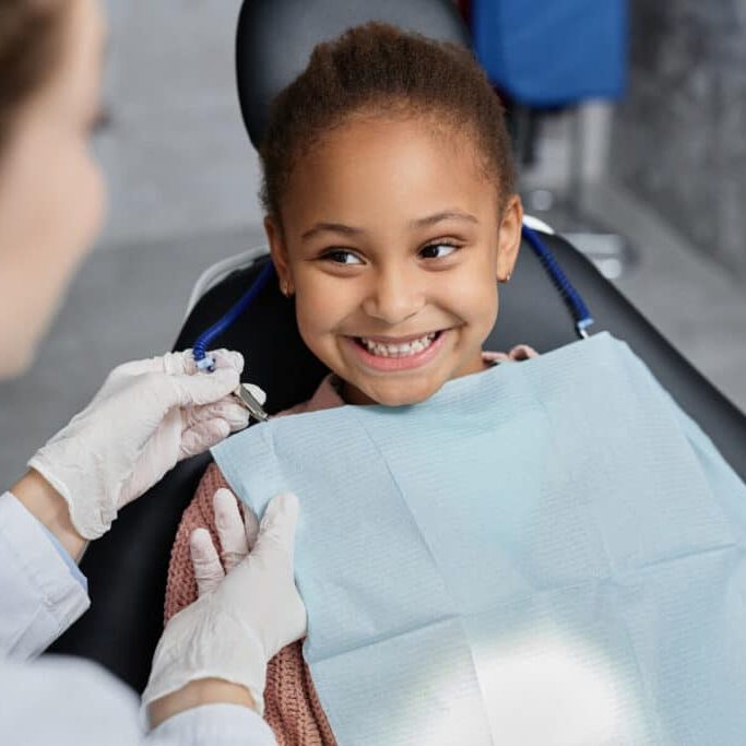 A smiling young girl sitting in a dental chair as a dentist in gloves prepares a bib.